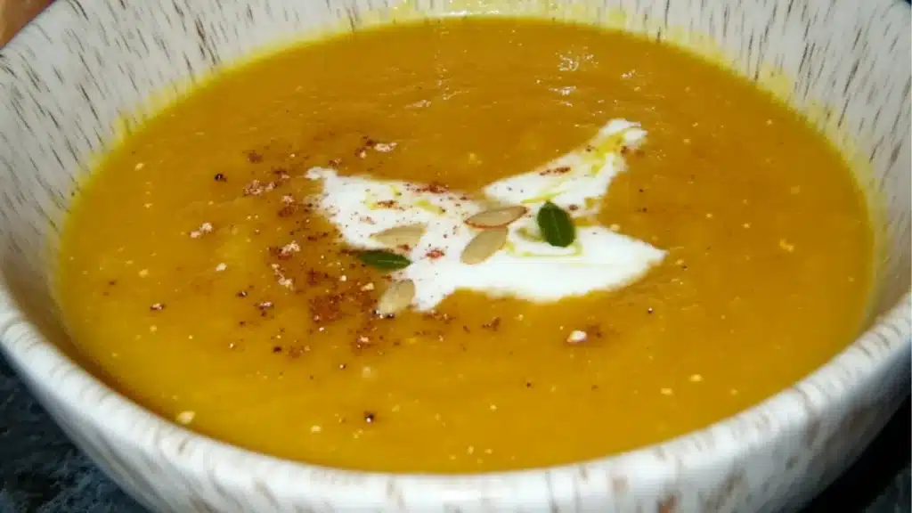 a white bowl filled with yellow soup and garnished with pumpkins on the side