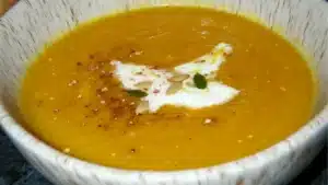 a white bowl filled with yellow soup and garnished with pumpkins on the side